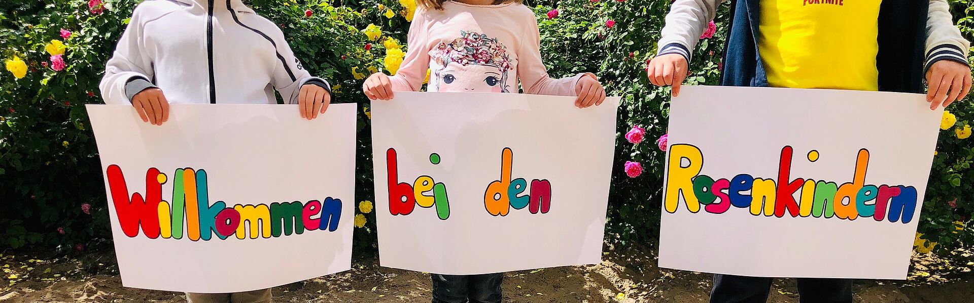 Drei Kinder stehen vor einer blühenden Rosenhecke im Sand und halten Schilder in den Händen, auf denen steht: "Willkommen bei den Rosenkindern"