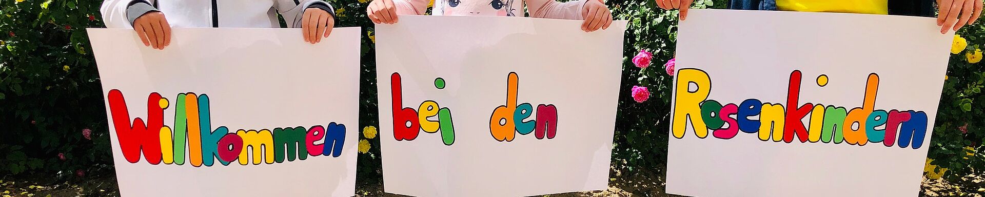 Drei Kinder stehen vor einer blühenden Rosenhecke im Sand und halten Schilder in den Händen, auf denen steht: "Willkommen bei den Rosenkindern"