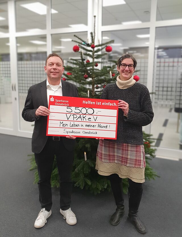 Ein Mann und eine Frau stehen mit goßem Scheck vor einem Weihnachtsbaum.
