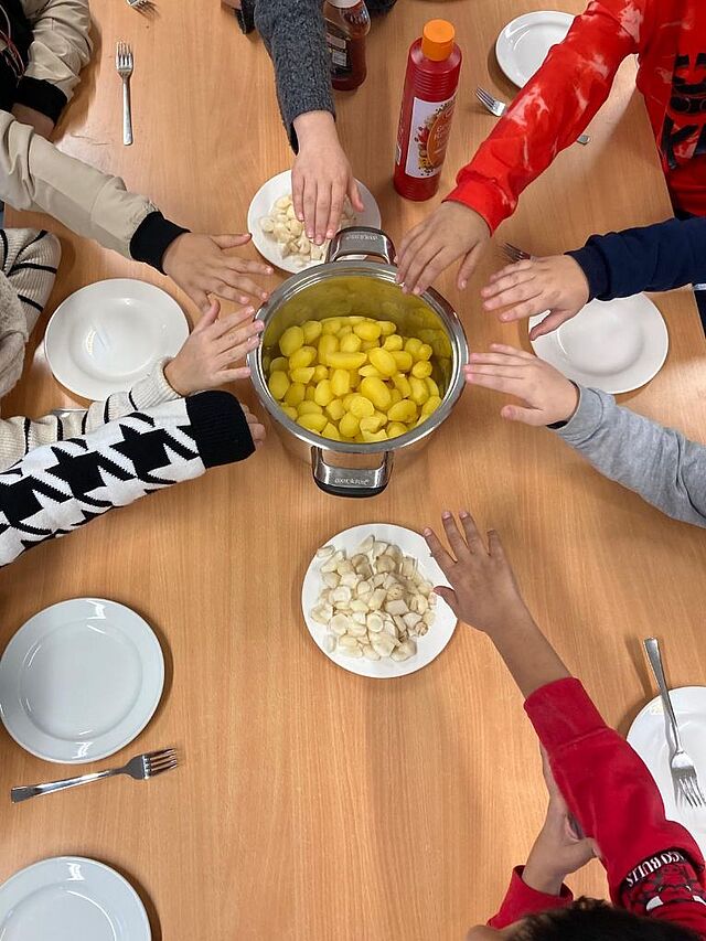 Auf dem Tisch steht ein Topf mit Kartoffeln. Mehrere Hände greifen nach dem Essen.