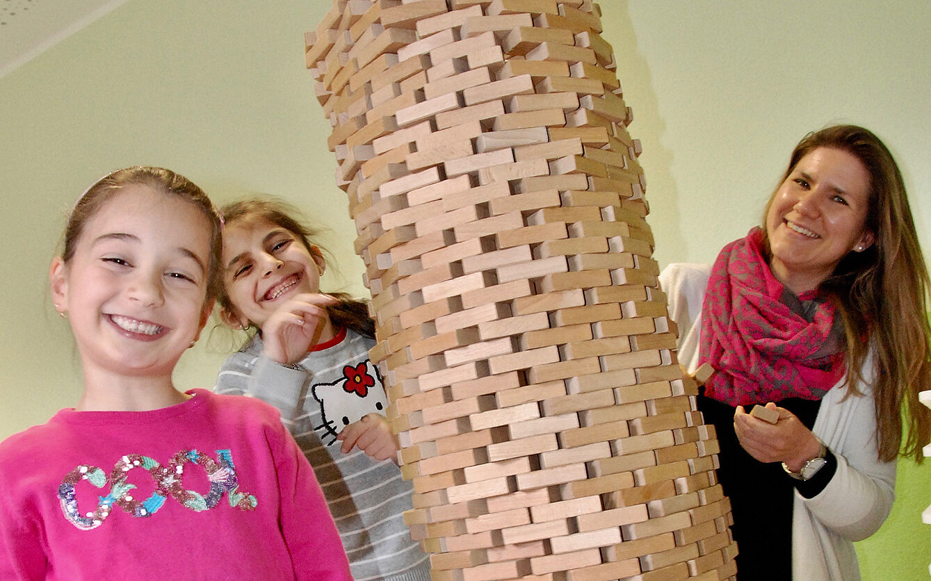 Zwei Mädchen und eine Erzieherin stehen an einem Turm aus JENGA-Klötzchen.