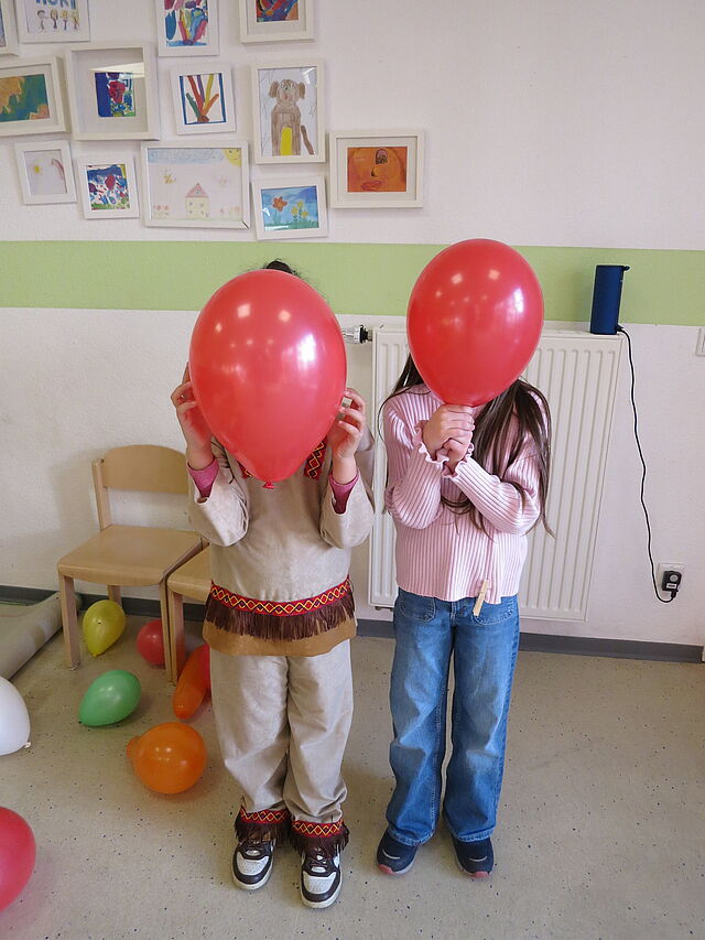Zwei verkleidete Kinder halten sich rote Luftballons vor das Gesicht.